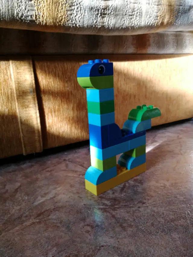 A simple long-necked dinosaur built from blue and green toy bricks, standing on a wooden floor