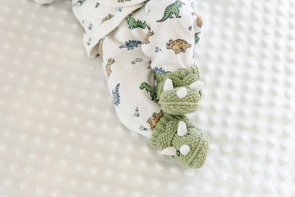 A baby's feet in green knitted triceratops booties, with a dinosaur-print sleeper, on a white dotted blanket