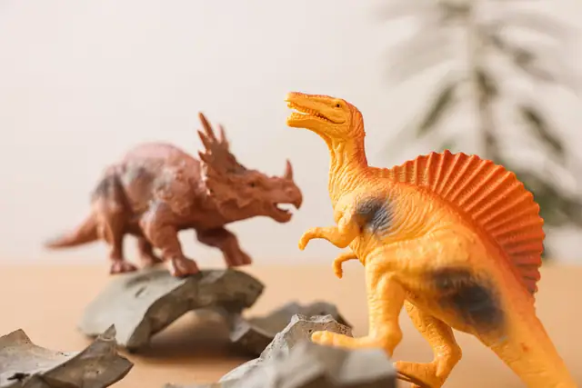 A horned dinosaur figure and an orange sail-backed dinosaur figure on a tabletop among broken shards