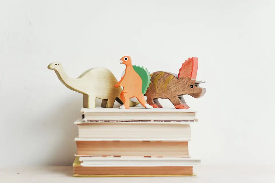 Three painted wooden dinosaur toys standing on a stack of hardcover books against a white wall